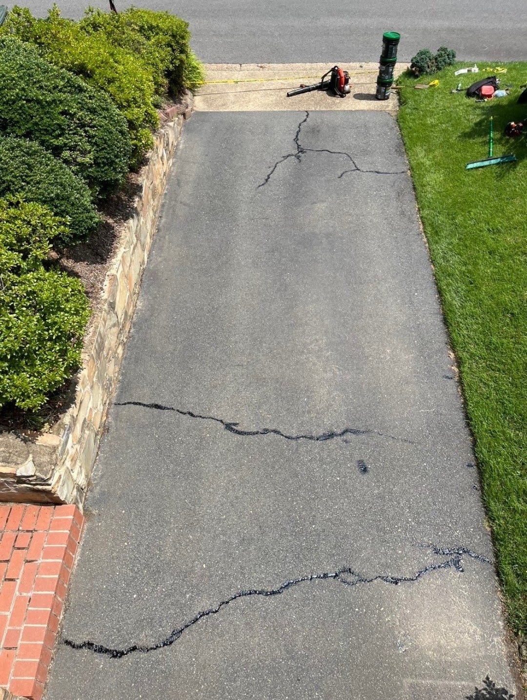 Aerial view of a cracked driveway with caulk lines, bordered by green grass and hedges, worker visible at top